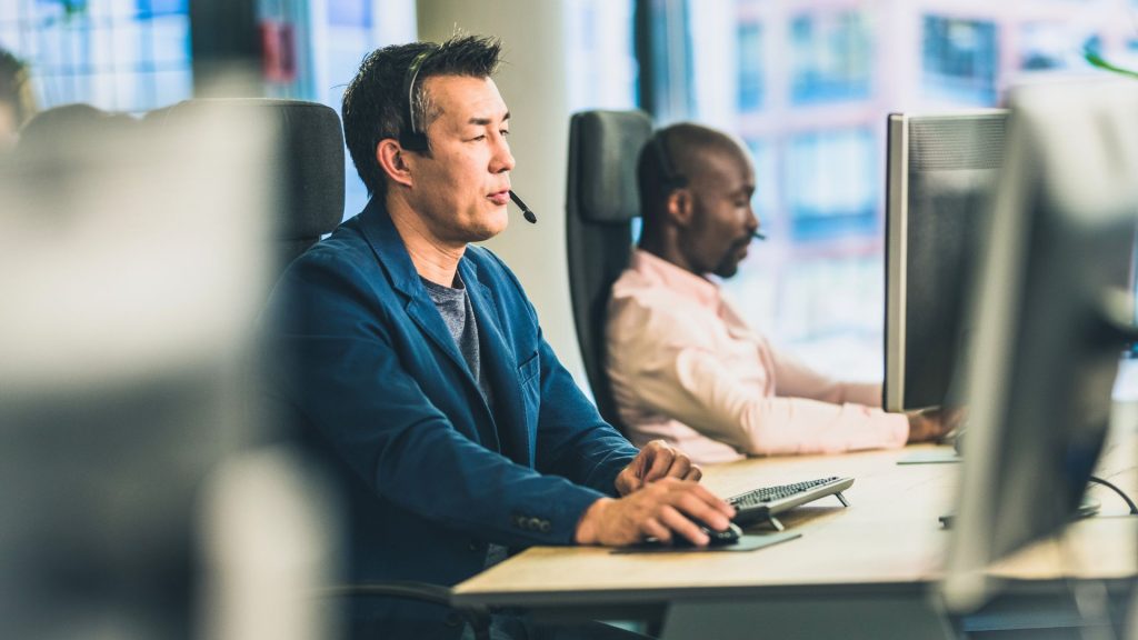 A man seated at a desk, focused on his computer screen, with papers and a pen nearby.